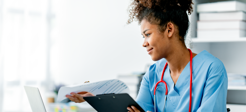 Female healthcare worker in blue scrubs reviews documents in a bright office.