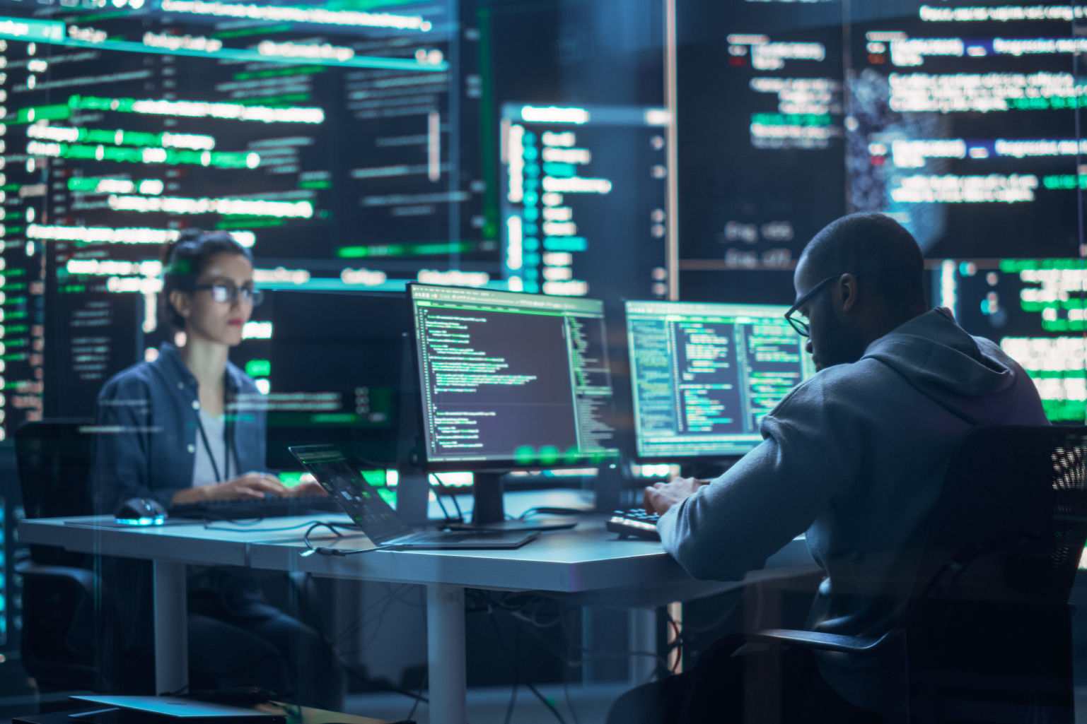 Two people working on computers with multiple monitors displaying code in a tech office.